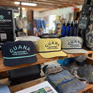 Three 'GUANA Outpost' hats displayed on a wooden shelf with various items in the background.