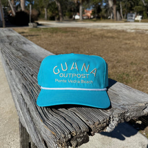 Blue cap with 'GUANA OUTPOST' text on a wooden bench outdoors
