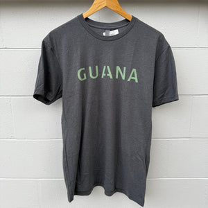 Gray t-shirt with 'GUANA' printed on it hanging on a wooden hanger against a light gray wall.