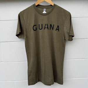 Olive green t-shirt with 'GUANA' text on a wooden hanger against a white wall.