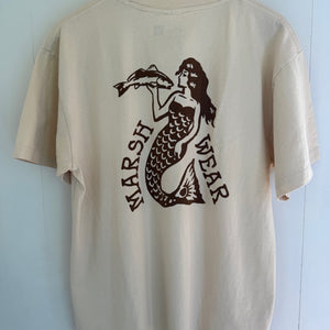 A cream-colored short-sleeve t-shirt hanging on a wooden hanger against a light blue paneled wall. The back of the shirt features a dark brown graphic of a mermaid holding a fish, with the text 'MARSH WEAR' printed vertically.
