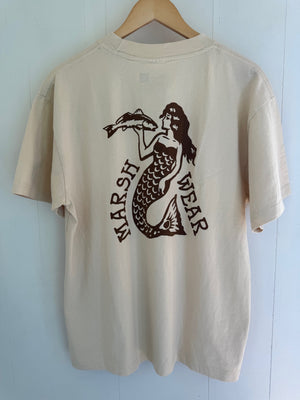 A cream-colored short-sleeve t-shirt hanging on a wooden hanger against a light blue paneled wall. The back of the shirt features a dark brown graphic of a mermaid holding a fish, with the text 'MARSH WEAR' printed vertically.