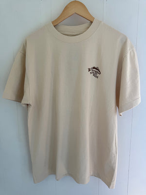 A beige short-sleeve crewneck t-shirt hanging on a wooden hanger against a white paneled wall. The shirt features a small dark brown fish graphic with the text 'MARSH WEAR' printed on the left chest.