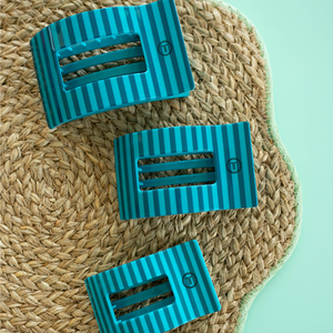 Three blue striped rectangular hair clips on a woven mat with a light green background